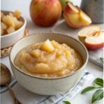 Bowl of warm homemade applesauce with cinnamon on rustic wooden board surrounded by fresh apples