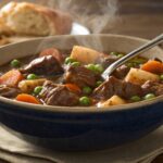 Bowl of slow cooker beef stew with tender beef chunks carrots potatoes peas rich brown gravy steam rising crusty bread side
